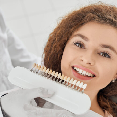 Closeup of beautiful young woman lying on dental chair, looking at camera and smiling while male dentist keeping in hands teeth color range. Girl doing whitening procedure in dental office.